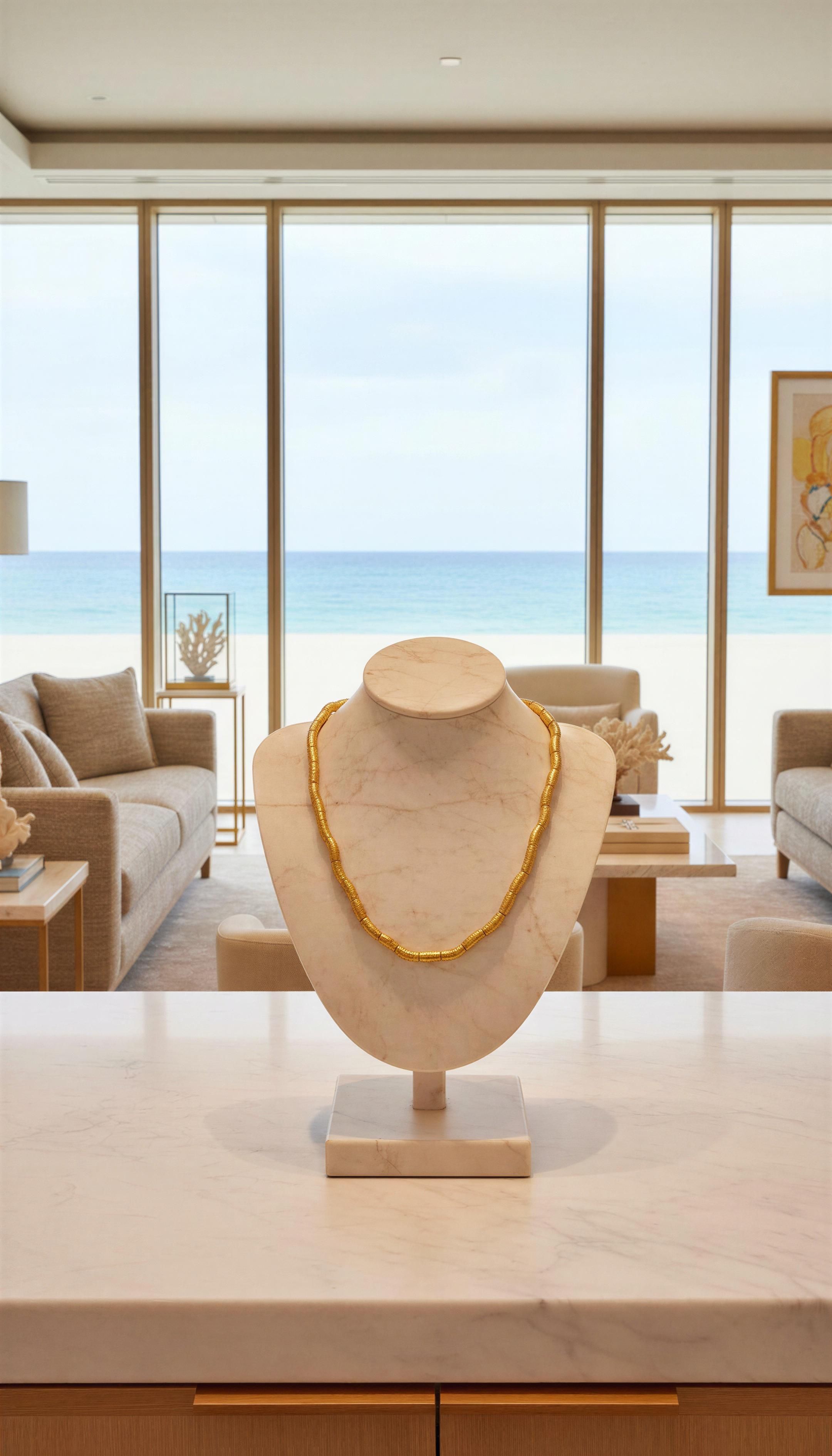 Elegant gold beaded necklace displayed on a marble bust in a bright, modern living room with an ocean view, showcasing Necklaces - AURELIS.