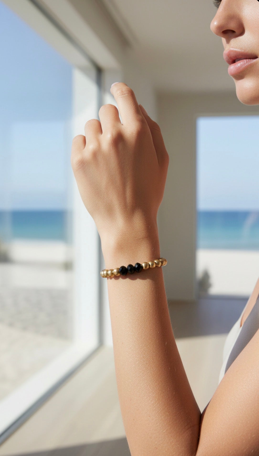 Close-up of a woman's wrist wearing the Limited Edition - Aurea Trinitas Antiqua gold and black beaded designer bracelet, with a serene ocean view in the background.