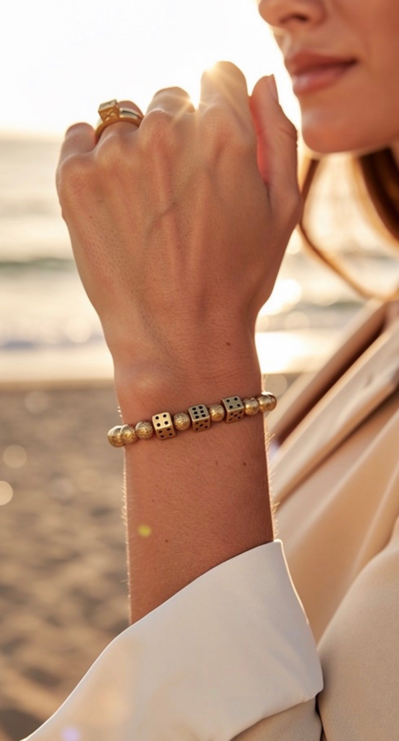 Close-up of a woman's wrist wearing a gold bead bracelet with dice-shaped charms, paired with a matching gold ring, captured in warm natural light near the beach, showcasing the Limited Edition - Alea Aurea Lux.