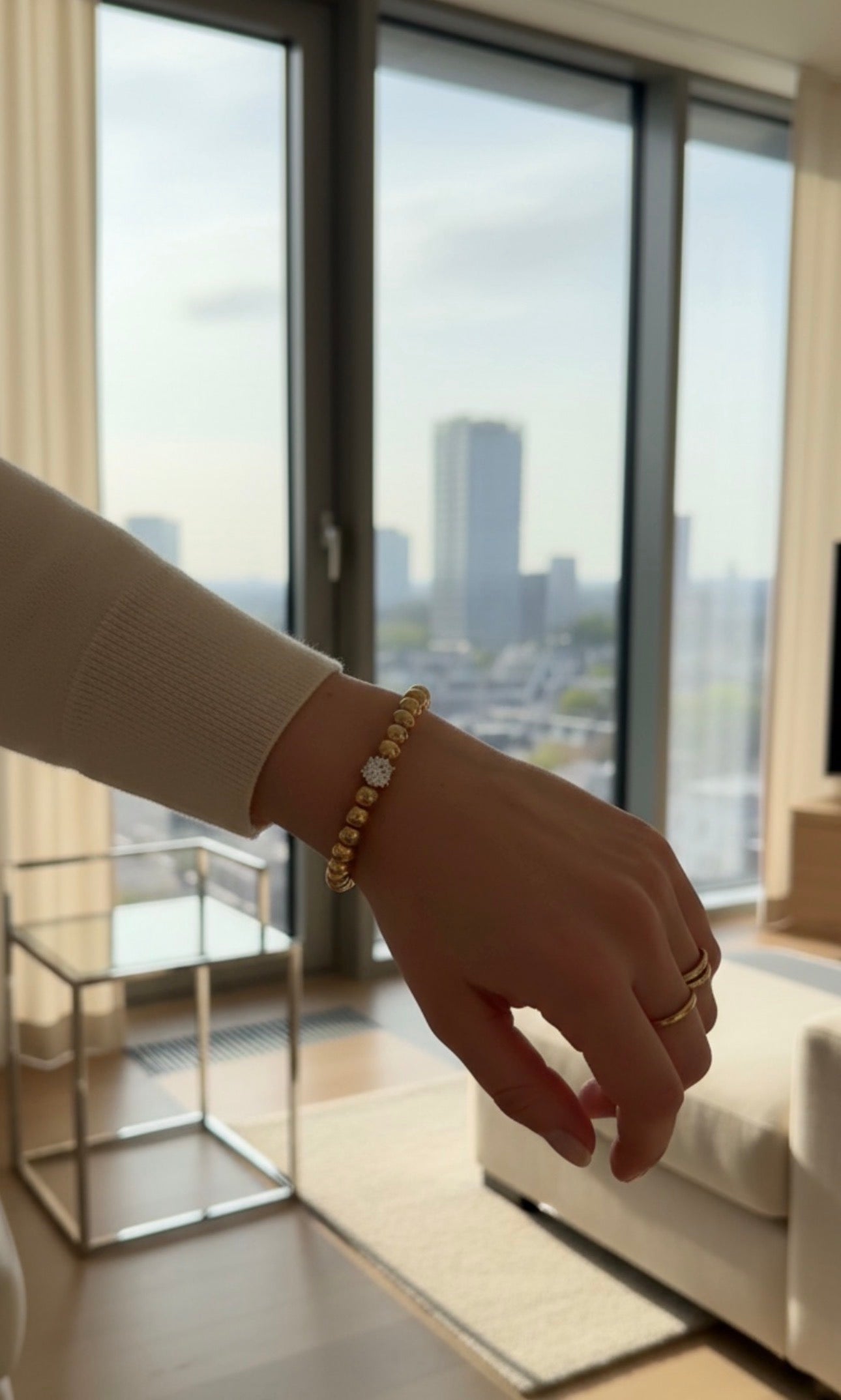 Close-up of a wrist adorned with a gold beaded bracelet featuring a central decorative bead, worn inside a modern living room with city views, showcasing The Timeless Collection β Aurea Claritas.
