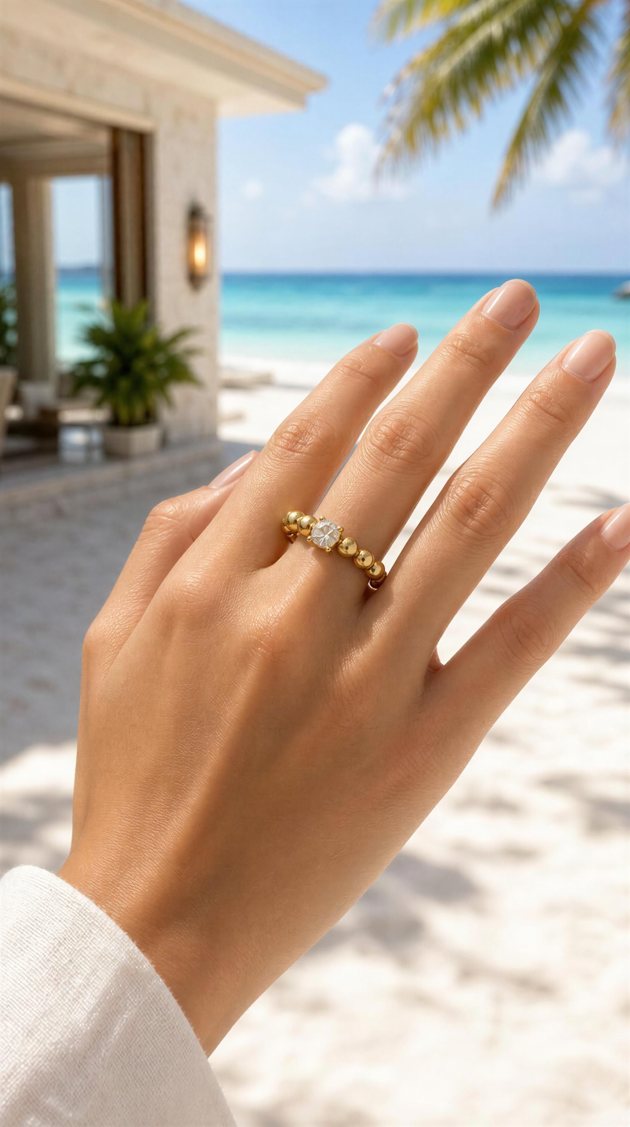 Hand wearing a RING - LUCENT ÉTOILE RING featuring gold beads and a central clear gemstone, with a serene beach and tropical palm background.