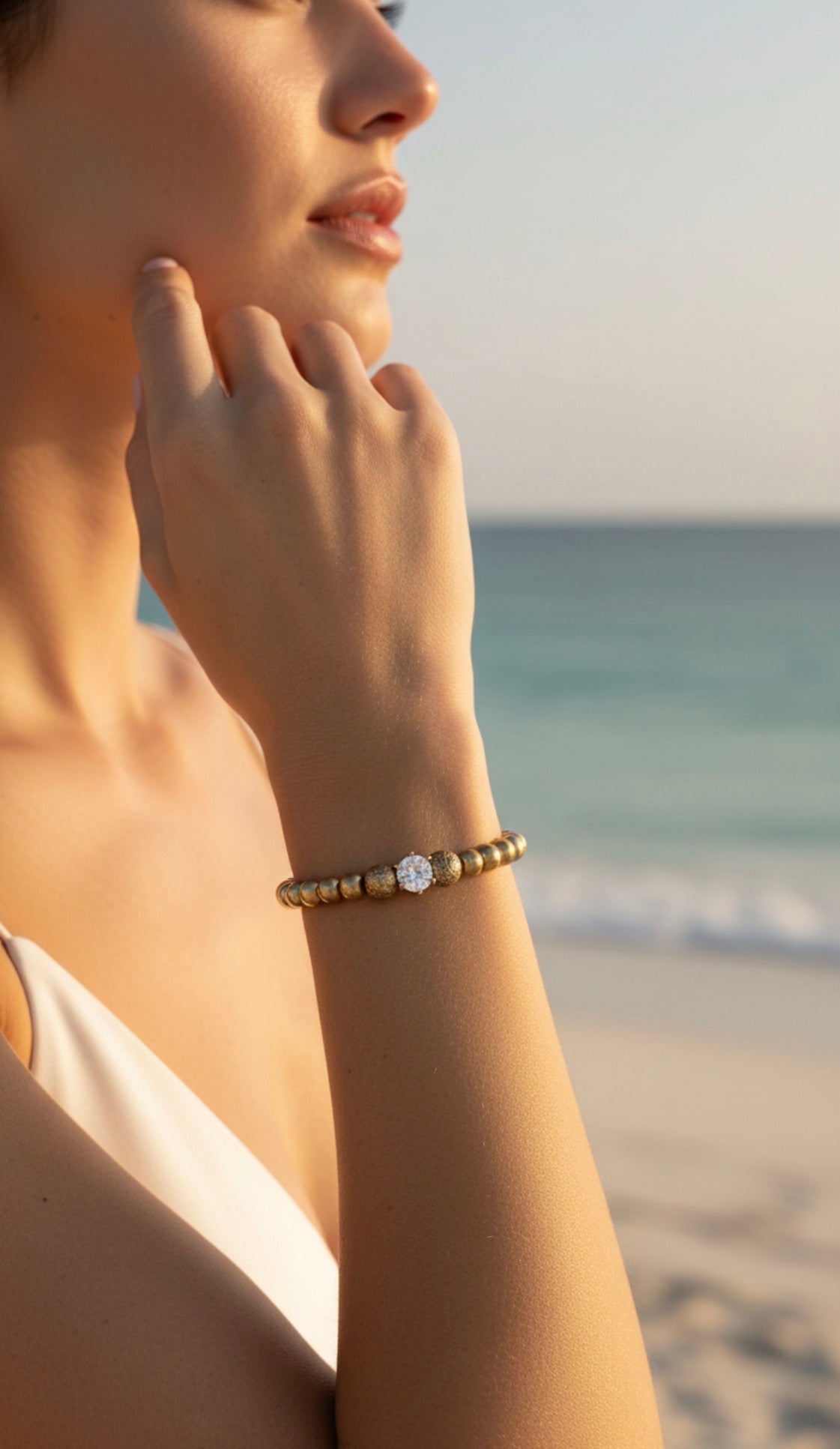 Close-up of a woman wearing a gold beaded bracelet with a central clear gemstone, showcasing the elegant Timeless Collection - Lux Textura by the ocean at sunset.