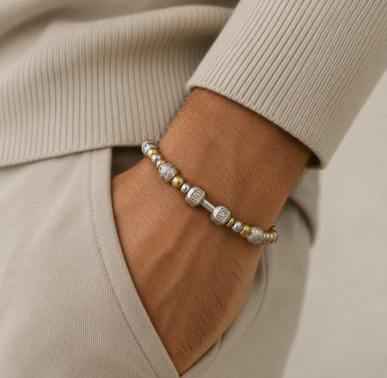 Close-up of a man's wrist wearing THE X FOR HIM - Live with Strength bracelet featuring mixed silver and gold beads with geometric and textured designs, styled with neutral beige clothing.
