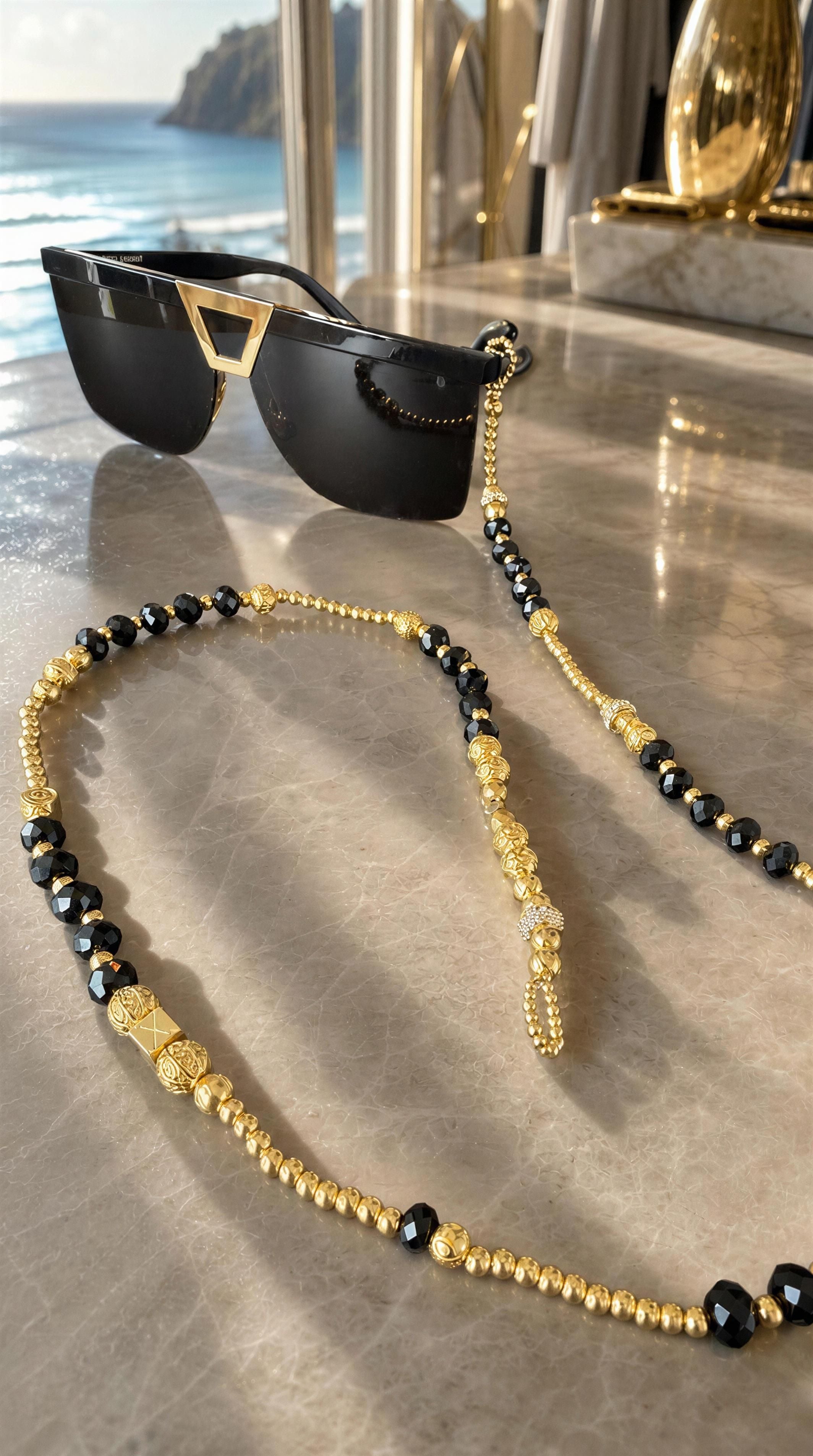 Black sunglasses with geometric gold accents paired with The Riviera Sunglass Chain - Noir Élégance featuring gold and black beads on a marble surface near a window overlooking the ocean.