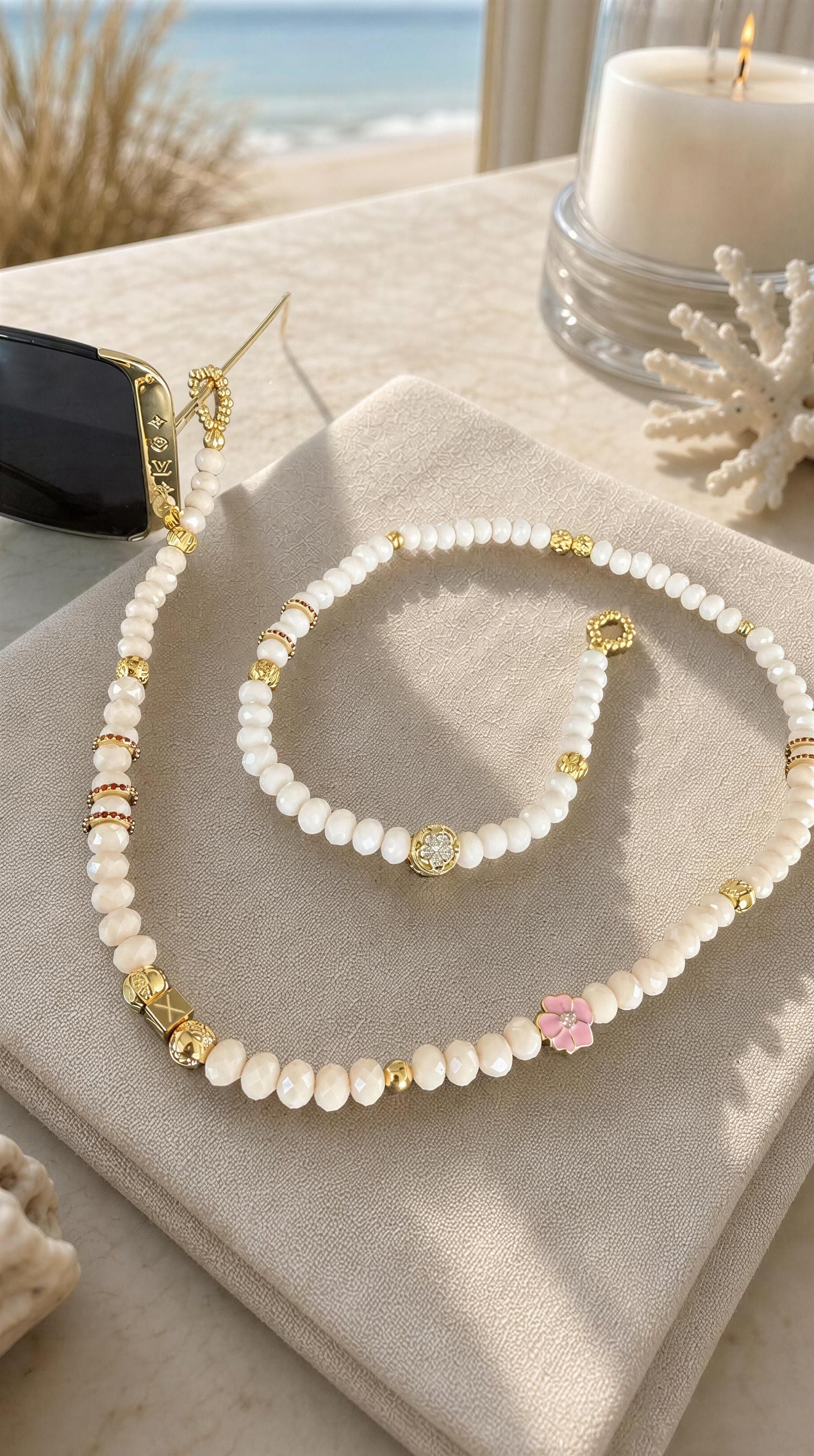 The Riviera Sunglass Chain – Rosé Lumière featuring delicate white beads accented with gold elements and a pink flower charm, styled elegantly with black sunglasses on a beige fabric.