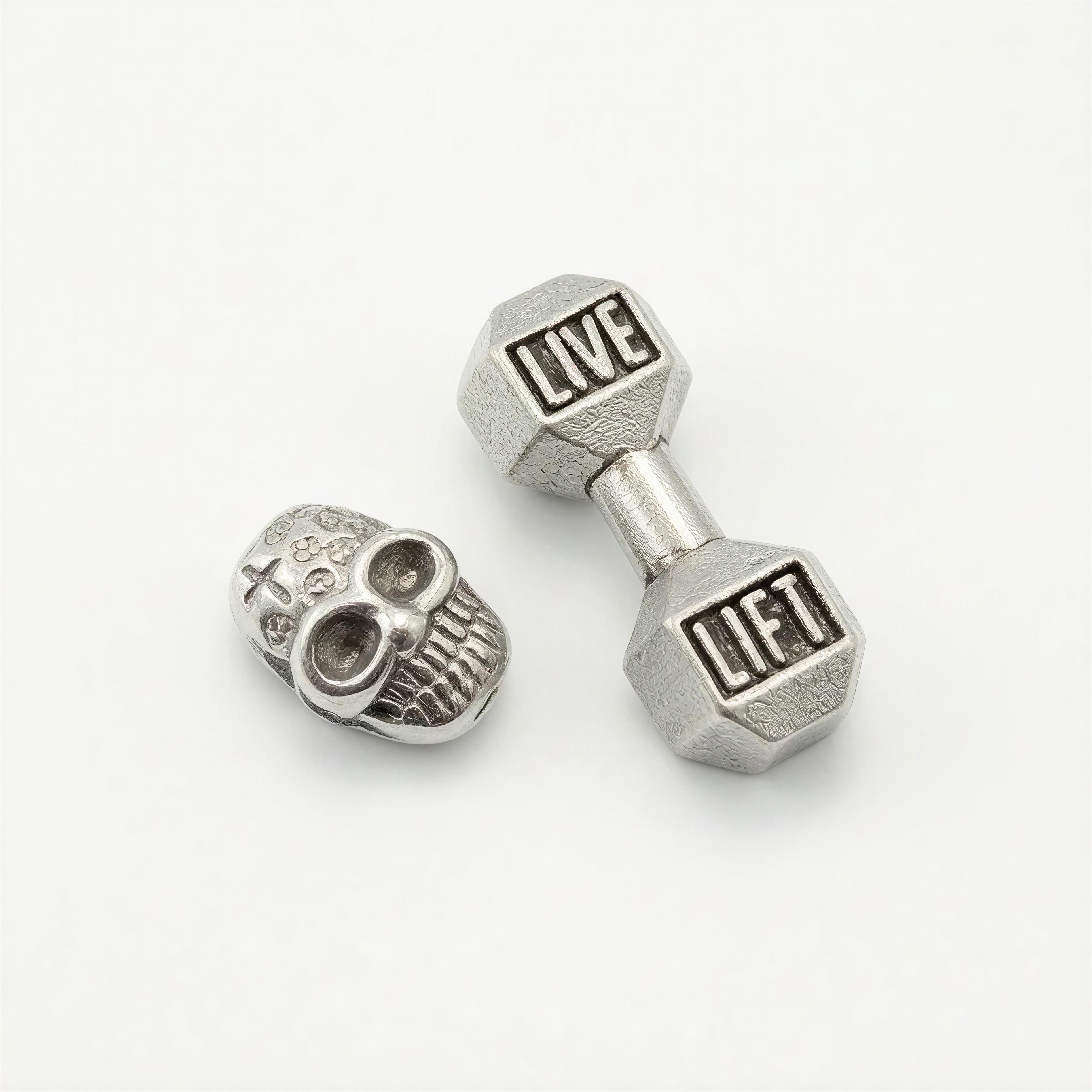 Two silver metallic charms on a white background, one shaped like a skull with intricate engravings and the other a dumbbell engraved with "LIVE" and "LIFT," representing THE X FOR HIM - Live with Strength.