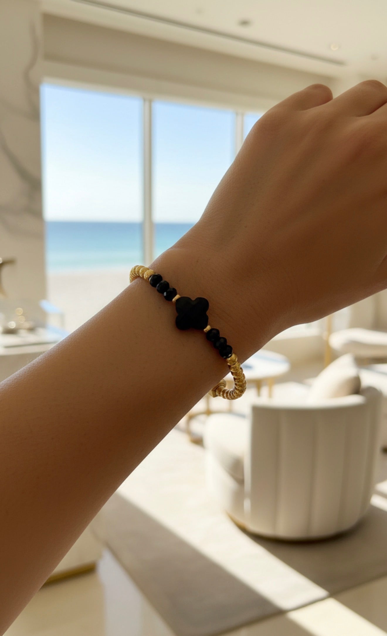 Delicate XX Élégance - Midnight Clover bracelet featuring black clover-shaped bead and gold accents, elegantly worn on wrist with a bright coastal view background.