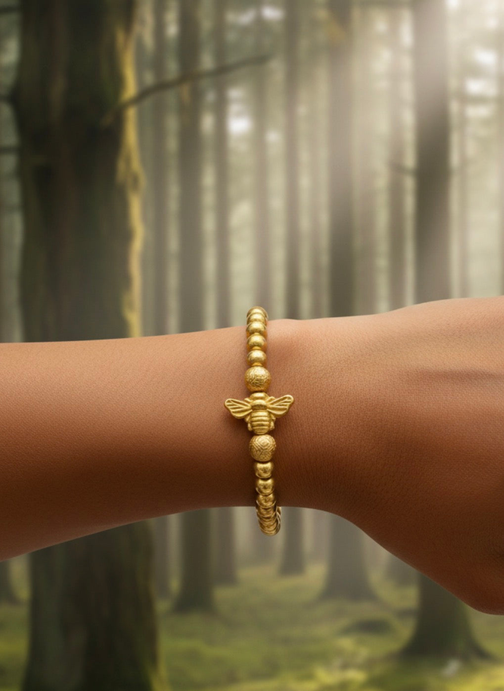 Close-up of a wrist wearing a gold beaded bracelet featuring a bee charm, set against a softly lit forest background, showcasing the Timeless Collection - Regina Apis.