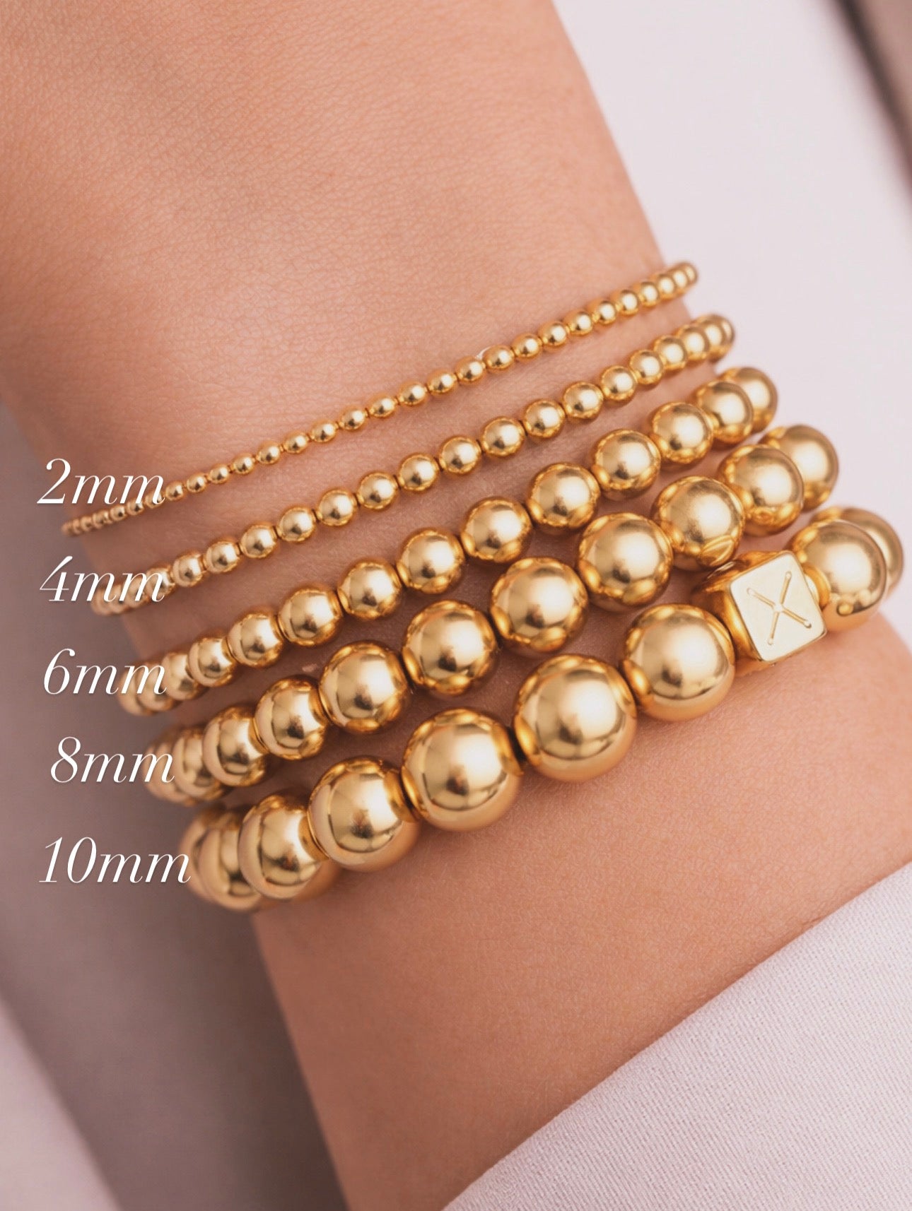 Stacked gold bead bracelets in varying sizes from 2mm to 10mm, featuring a designer 8mm bracelet from The Essential Collection - 8mm with a small cube bead engraved with an "X" for Atelier X luxury handcrafted jewelry.