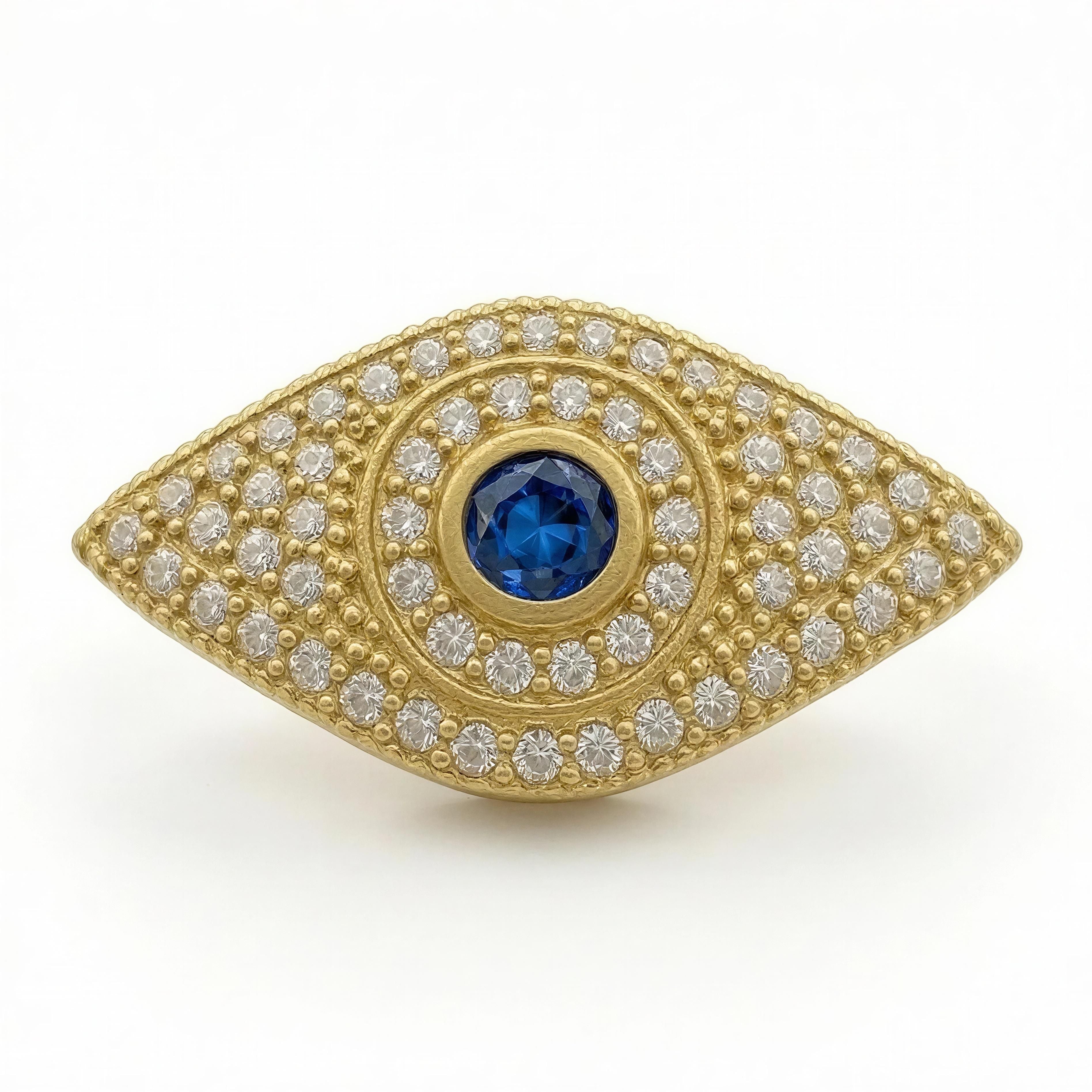 XX Élégance - Ojo Celestial features a luxurious gold eye-shaped design adorned with sparkling white stones and a vibrant blue gemstone centerpiece, embodying handcrafted elegance.